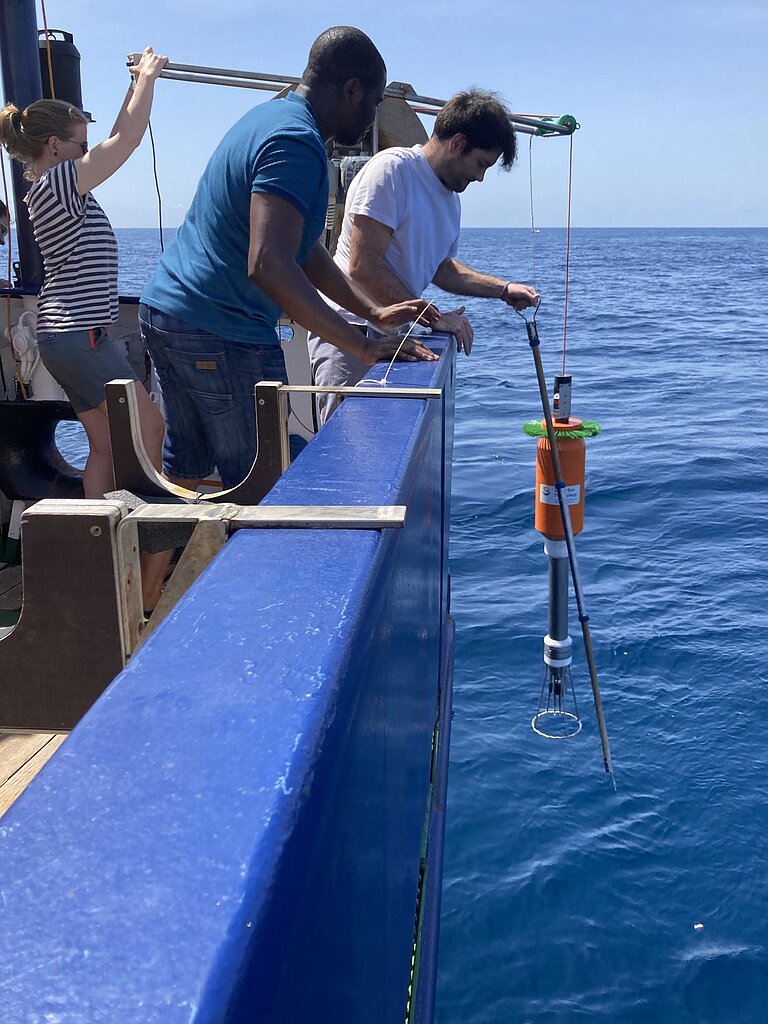 A woman and two men jointly lower a measuring device over the railing of a ship