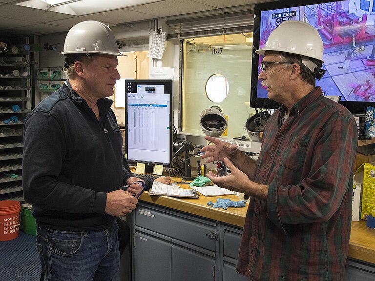 Zwei Männer mit Sicherheitshelmen stehen vor einer Arbeitsplatte in einer Werkstatt, über der ein großer Monitor hängt.