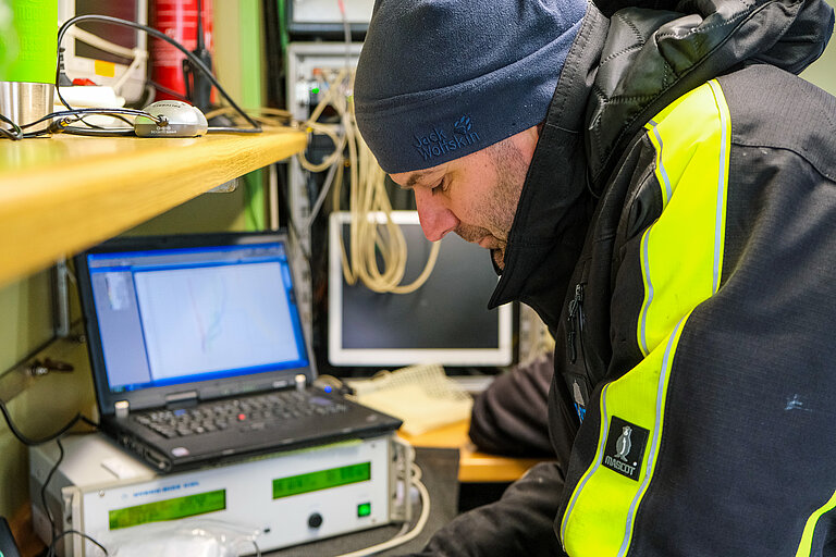 Ein Mann in warmer Kleidung arbeitet an einem Schreibtisch neben Messgeräten und einem aufgeklappten Laptop