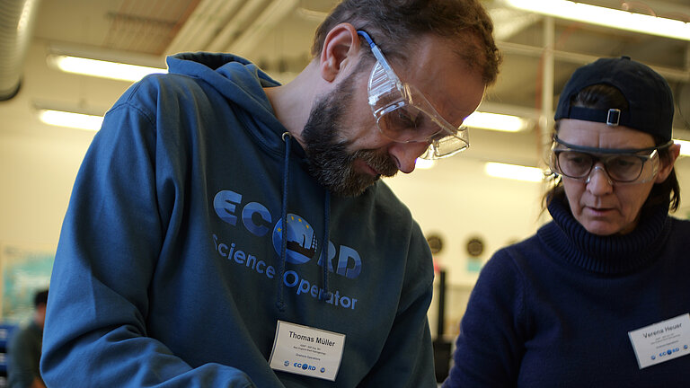 Zwei Personen, ein Mann und eine Frau. Der Mann trägt eine transparente Labor-Schutzbrille und bearbeitet auf dem Tisch liegendes Gestein mit einem Meißel und einem Kunststoffhammer