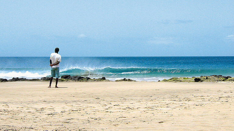 Eine Person schaut vom Strand aus auf das Meer, wo sich eine Welle bricht.