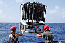 Ein Mann und eine Frau händeln an einer Bordwand stehen einen Kranzwasserschöpfer. 
