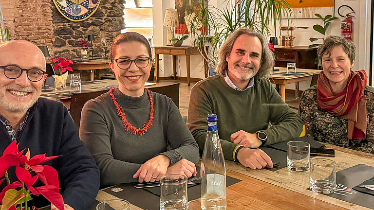 Zwei Männer und zwei Frauen sitzen gemeinsam am Tisch und lächeln in die Kamera.