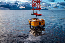 Ein gelber eckiger Tauchroboter wird an einem roten Kran ins Wasser gelassen.