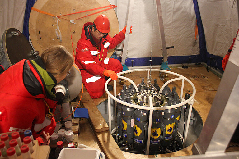 Ein Forscher und eine Forscherin in dicken roten Anzügen in einem Zelt an einem Loch, in das ein wissenschaftliches Messgerät hinuntergelassen wird