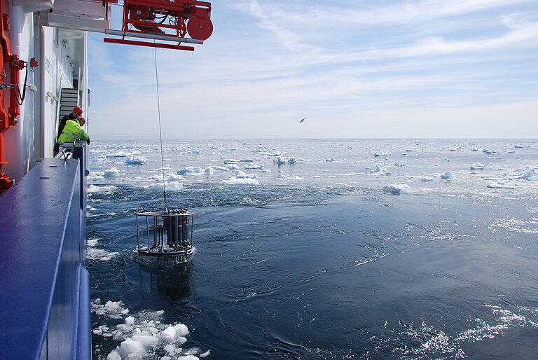 Ein Kranzwasserschöpfer wird von Bord eines Forschungsschiffes in das Meer gelassen. Auf dem Wasser sind Eisschollen zu sehen.