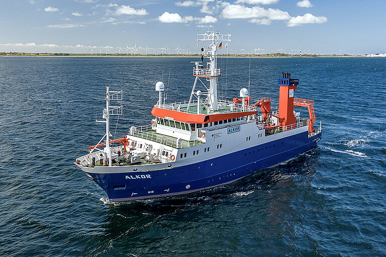 Ein mittelgroßes Forschungsschiff fährt auf dem Meer unweit der Küste, wo Windkraftanlagen zu sehen sind