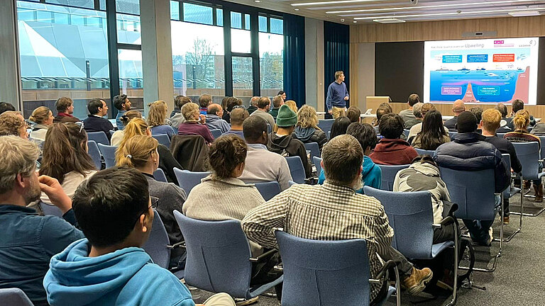 Ein Konferenzsaal mit Publikum, vorne an der Leinwand steht ein Mann und spricht