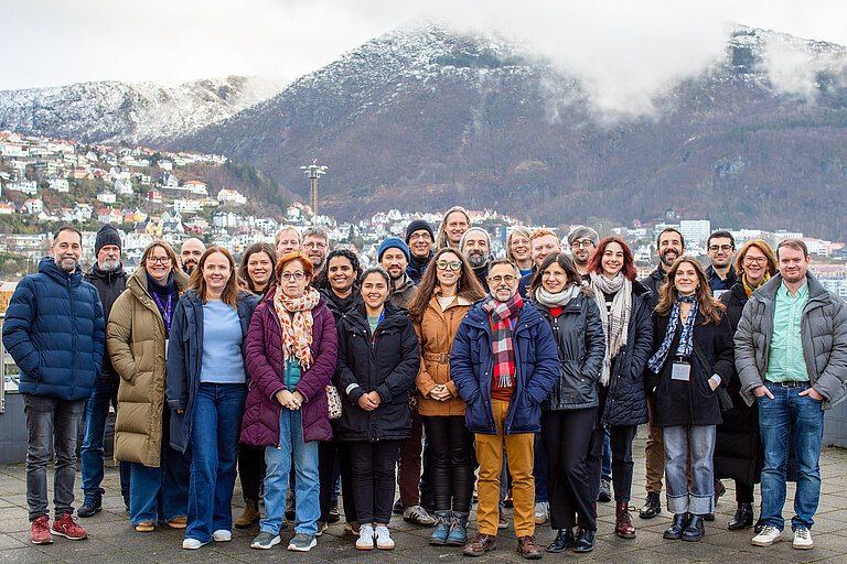 Eine Gruppe von etwa 25 Menschen in Winterjacken steht vor bebauten Hängen und Bergen, auf deren Spitzen Schnee liegt 