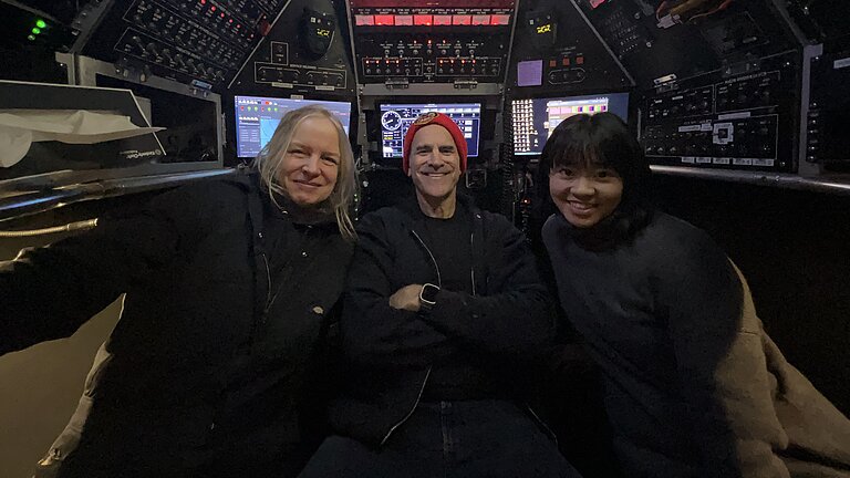 Three people sit in the ALVIN submersible. Buttons and displays all around them. They laugh into the camera.