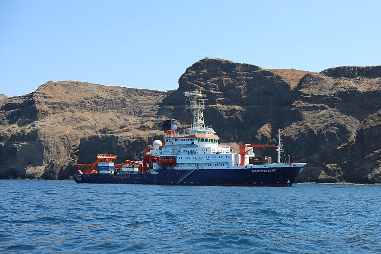 Das Forschungsschiff METEOR auf blauem Wasser, im Hintergrund grünlich-braune Felsen