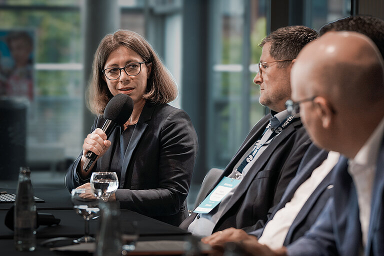 Eine Frau und mehrere Männer in blauen Anzügen sitzen hinter einem langen Tisch, die Frau spricht in ein Mikrophon