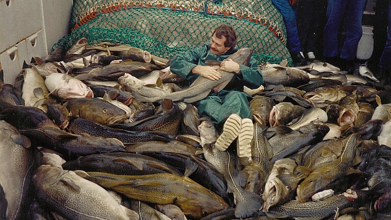Ein Mann in einem grünen wasserdichten Overall liegt auf einem Berg riesiger Fische und hält ein Exemplar im Arm, das über einen Meter lang ist
