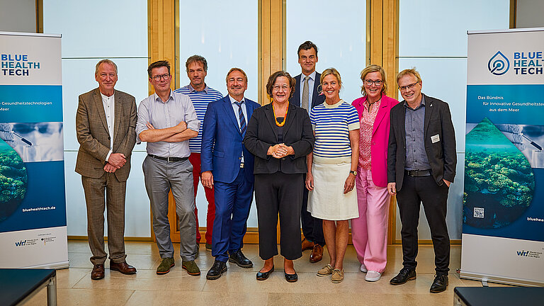 Eine Gruppe von Männern und Frauen in sommerlicher Kleidung, rechts und links stehen Banner, auf denen BlueHealth zu lesen ist.