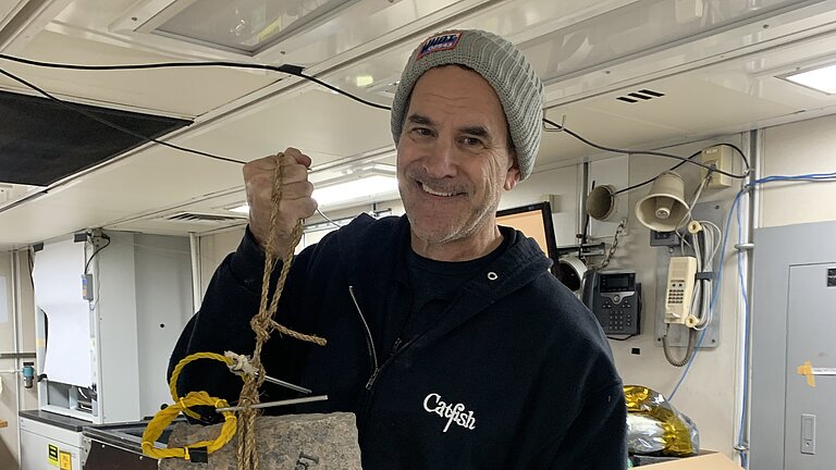 Alvin pilot Bruce Strickrott prepares to deploy the memorial stone with the submersible Alvin. He stands at a table and holds the stone up to the camera.