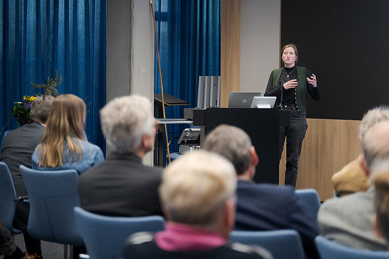 Eine junge Frau steht an einem Rednerpult und spricht zu Publikum
