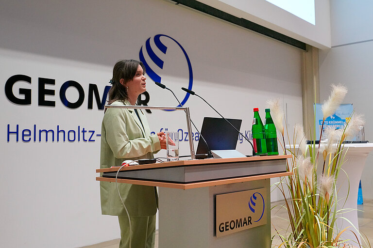 Eine junge Frau mit dunklen Haaren und grünem Hosenanzug steht an einem Stehpult und hält einen Vortrag