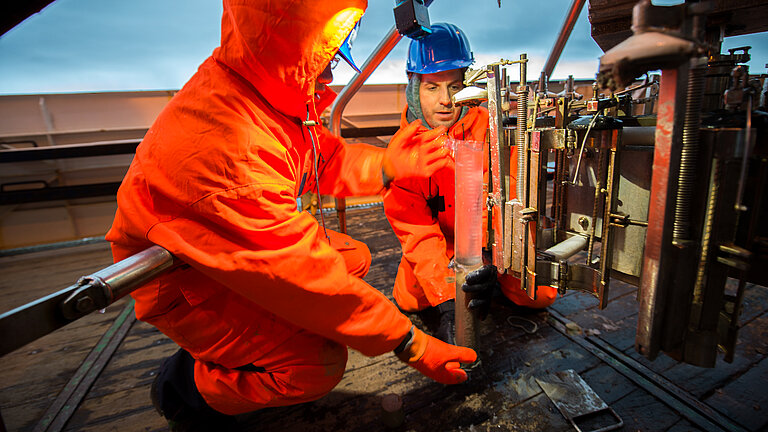  Two men in bright orange overalls aboard a ship, between them a transparent tube, each half filled with sediment and water.