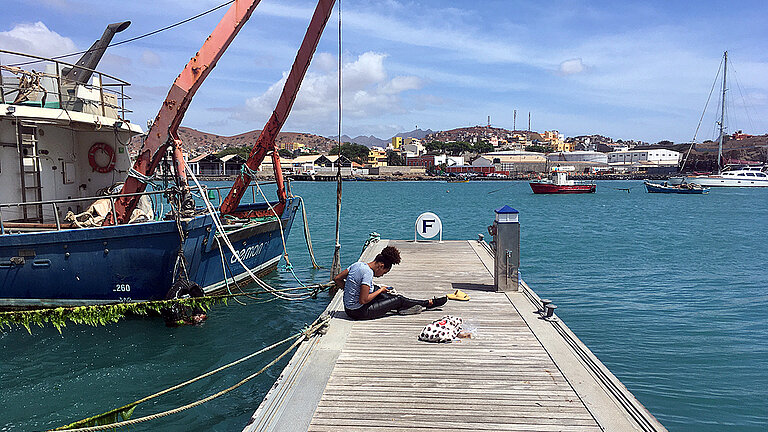 Ein Steg ragt in türkisblaues Wasser, darauf sitzt eine junge Frau 