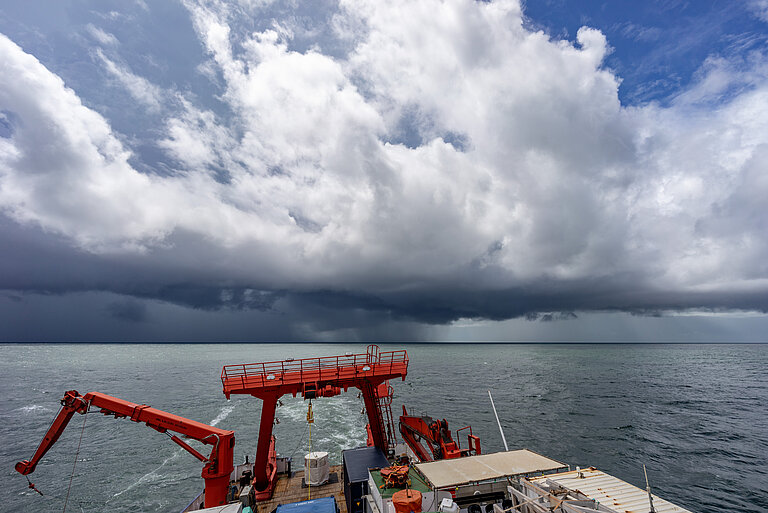 Dramatische Wolken türmen sich am Himmel über dem Meer, im Vordergrund ist der Bug eines Forschungsschiffes zu sehen.