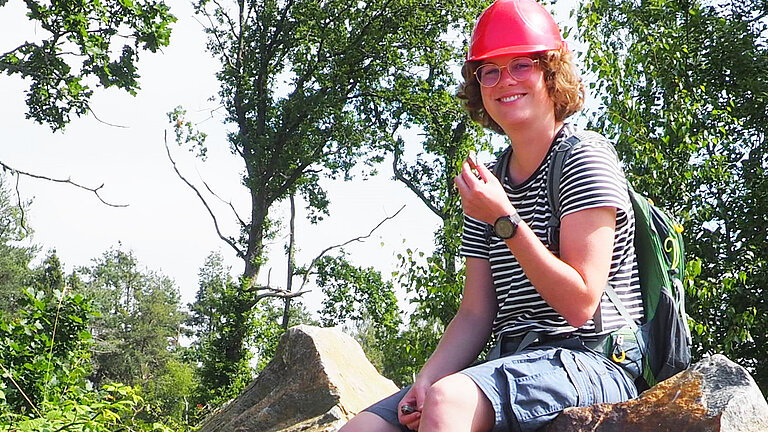 A young woman with a red hard hat sits on a large stone