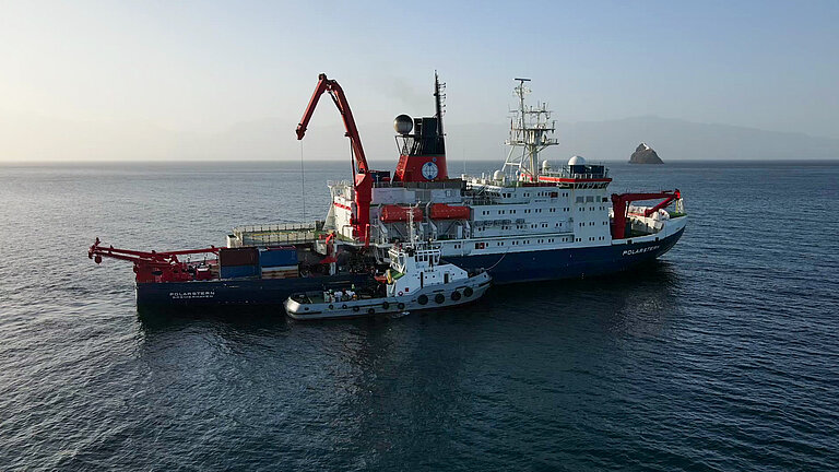 Ein großes Forschungsschiff mit einem markanten roten Kran liegt in ruhiger See auf Reede, ein kleines Schiff hat quer dazu festgemacht.