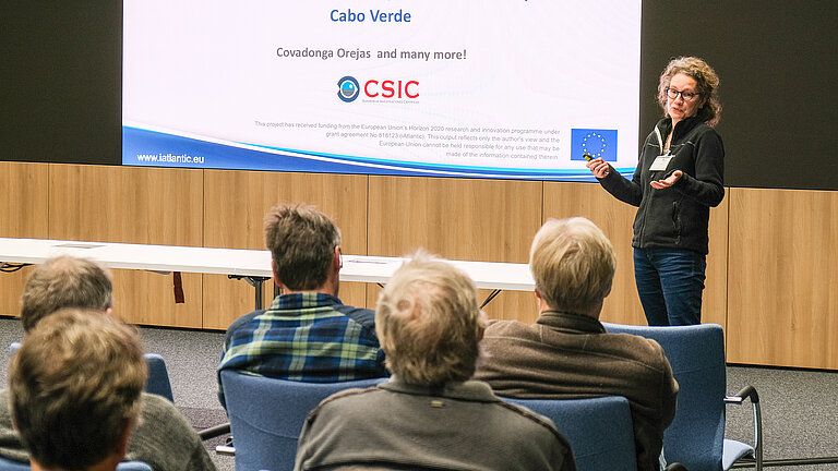 Eine Frau steht vor einer Leinwand und spricht zu einem Auditorium.