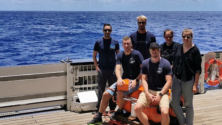 Sechs Männer und Frauen in T-Shirts sitzen an Deck eines Schiffes