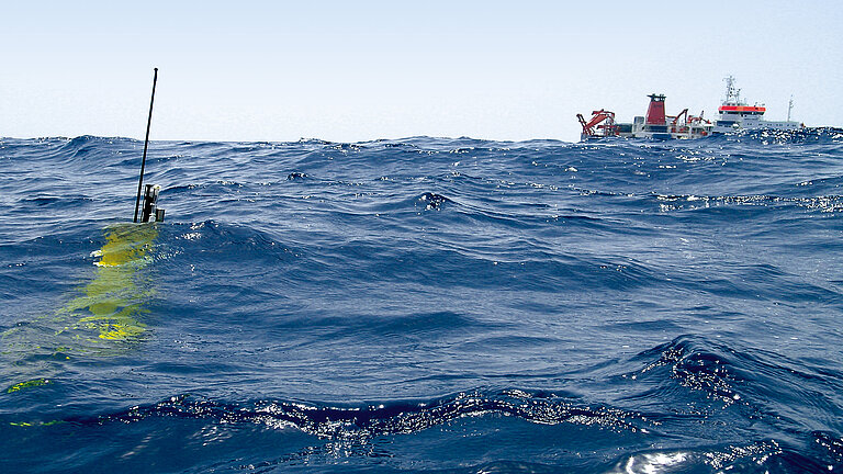 Eine gelbe Messboje im Wasser, am Horizont ein Forschungsschiff