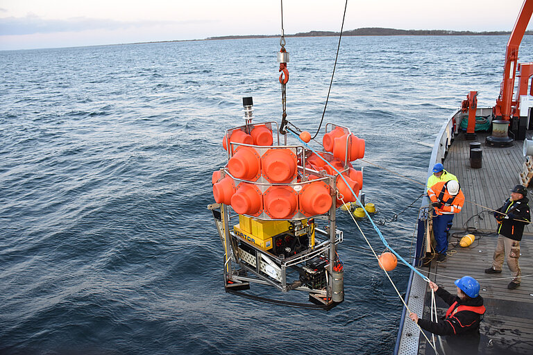 A huge instrument with orange bubbles is lifted over the edge of the ship by a crane. People stand by and watch.