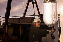 Ein Mitarbeiter auf einem Forschungschiff mit weißem Sicherheitshelm führt einen mit Wasser befüllten gläsernen Behälter, der an einem Gurt hängt, vorsichtig zurück an Bord des Schiffes.