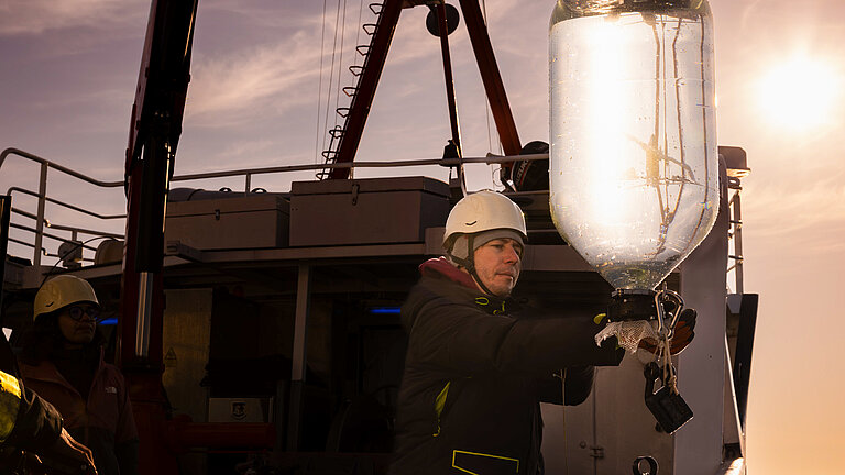  An employee on a research vessel wearing a white safety helmet carefully guides a glass container filled with water, which is hanging from a belt, back on board the ship.