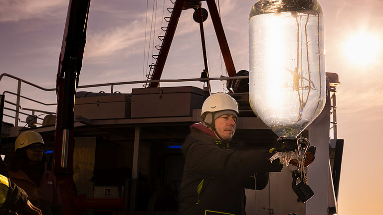 Ein Mitarbeiter auf einem Forschungschiff mit weißem Sicherheitshelm führt einen mit Wasser befüllten gläsernen Behälter, der an einem Gurt hängt, vorsichtig zurück an Bord des Schiffes.