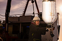  An employee on a research vessel wearing a white safety helmet carefully guides a glass container filled with water, which is hanging from a belt, back on board the ship.
