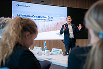 A man in a suit speaks in front of a large screen