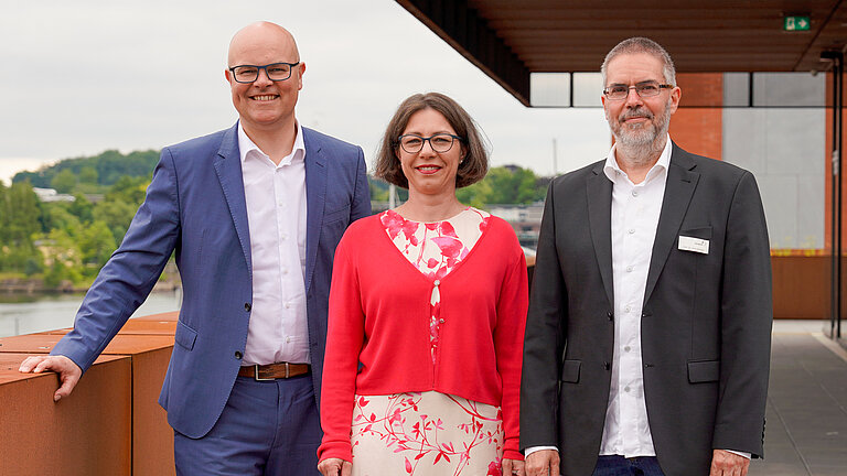 Zwei Männer und eine Frau stehen auf einer Dachterrasse und blicken in die Kamera