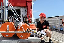 Frau mit roten Schutzhelm und Klemmbrett in der Hand hockt auf einem Schiffsdeck vor einem Gerät.  