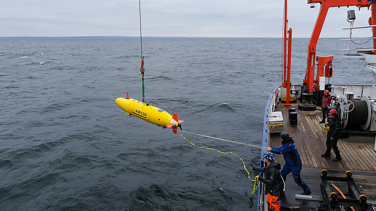 Graues Meer mit Wellen, darauf ein Schiff mit roten Aufbauten. An Bord stehen Männer mit Schutzhelmen, zwei von ihnen haben gelbe Leinen in der Hand, mit dem sie ein am Kran hängendes zigarrenförmiges gelbes Unterwasserfahrzeug  dirigieren.