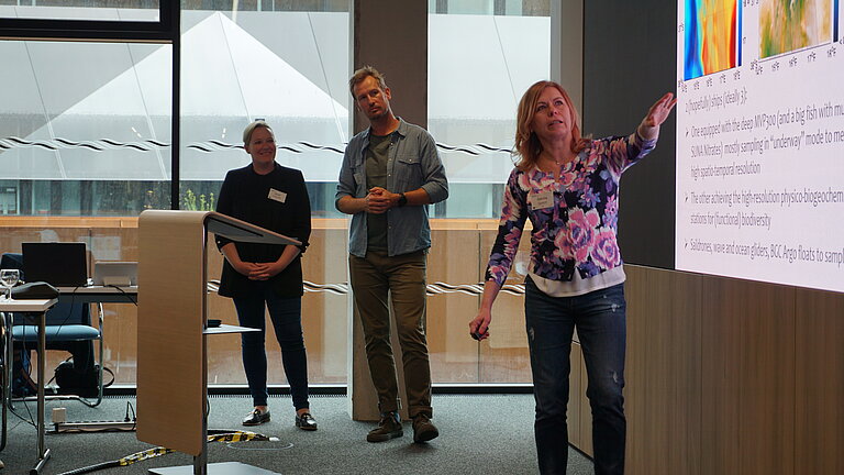 Drei Personen stehen vor einem Bildschirm und präsentieren etwas.