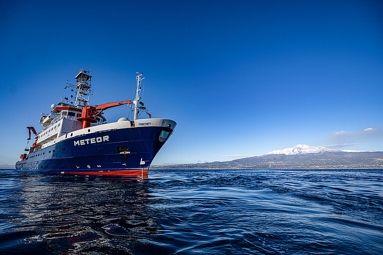 Ein Schiff auf blauem Meer, im Hintergrund die Küste mit einem Vulkan