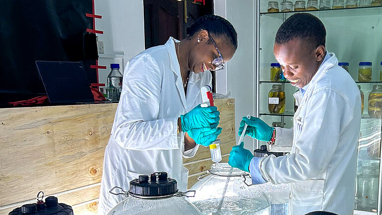 Drei dunkelhäutige Personen mit weißen Laborkitteln arbeiten in einem Labor an großen durchsichtigen, mit Wasser befüllten gläsernen Behältern