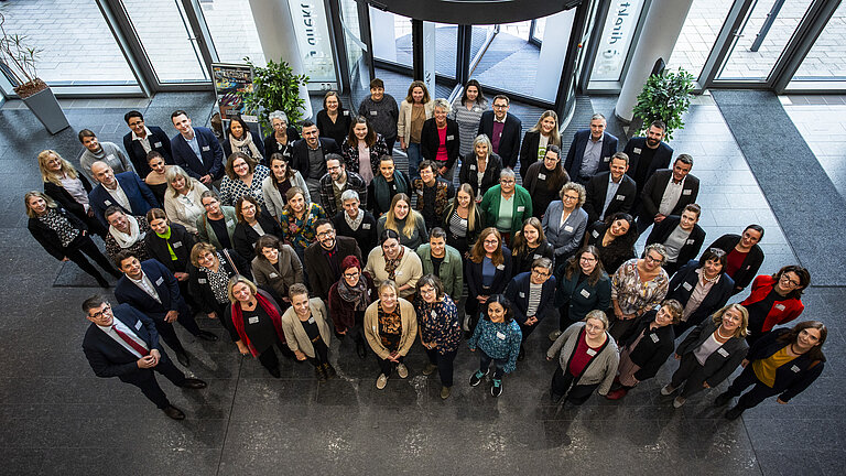 Eine Gruppe Menschen steht im Foyer eines Geschäftshauses und blickt in die Kamera über ihnen