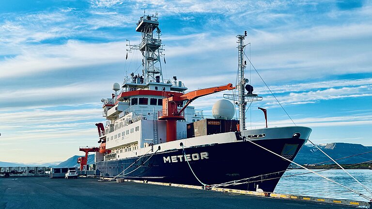 Das Forschungsschiff METEOR liegt im Hafen. Der Himmel ist blau mit Wolken. 