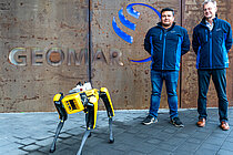 A yellow robot dog stands in front of a building with the word "GEOMAR" on it, with two men in blue jackets behind it.