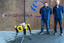 Ein gelber Roboterhund steht vor einem Gebäude mit dem Schriftzug "GEOMAR", dahinter zwei Männer in blauen Jacken