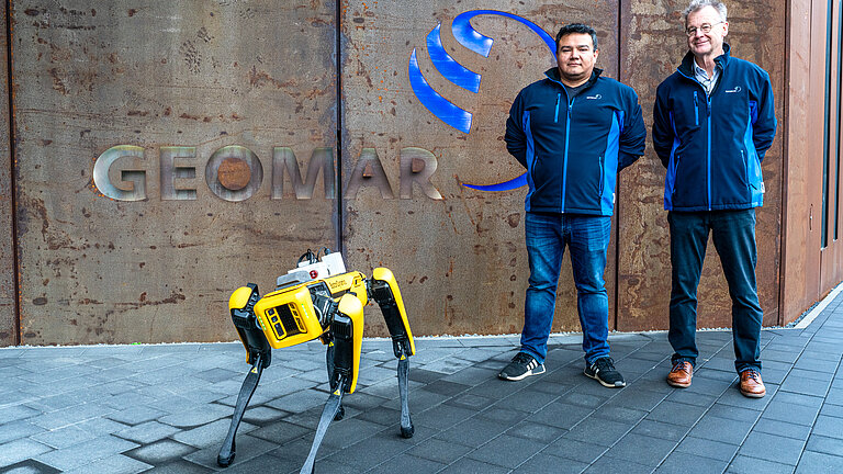 A yellow robot dog stands in front of a building with the word "GEOMAR" on it, with two men in blue jackets behind it.