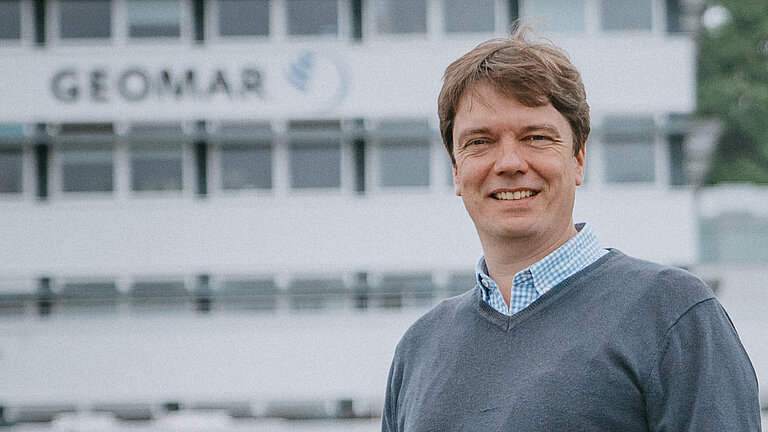 Ein Mann im grauen Pullover vor einem großen weißen Gebäude mit der Aufschrift GEOMAR