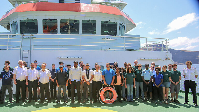 Schiffscrew in weißen Hemden und schwarzen Hosen und wissenschaftliche Crew und Besucher posieren auf dem Helikopterdeck eines Forschungsschiffes