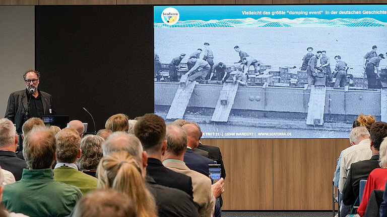 Ein Mann spricht vor Publikum in einem Konferenzraum. Auf der Leinwand ist ein Schwarz-Weiß-Foto zu sehen, auf dem Munitionskisten von Bord eines Schiffes ins Meer geworfen werden.