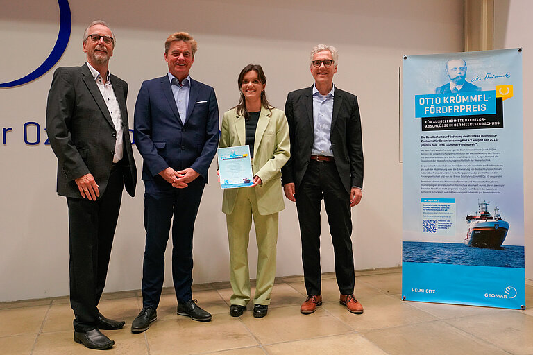 Eine junge Frau in hellgrünem Hosenanzug und drei ältere Herren im Anzug in einem Hörsaal